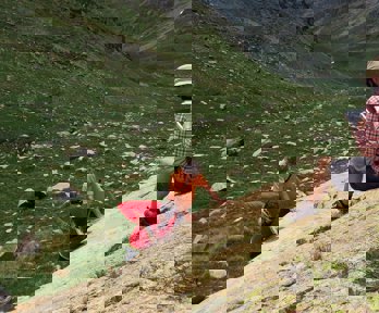 A researcher sitting on a rocky slope in a valley writing in a notebook while two others stand testing the rock hardness.