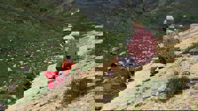 A researcher sitting on a rocky slope in a valley writing in a notebook while two others stand testing the rock hardness.