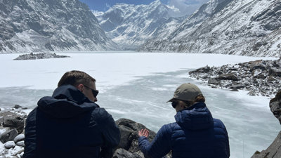 Two people talking, sat by a frozen lake with snow-capped mountains in the background.