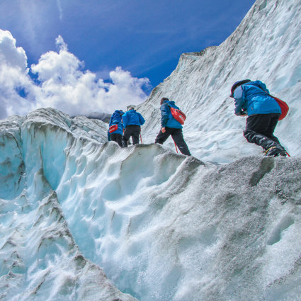 A group of people, all wearing blue jackets and carrying red backpacks, are walking up the side of a glacier. The glacier is tinted blue, and the edges are grey.