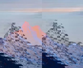 A mountain in the early or late sun, with its tip in soft sunlight. The mountain is purple, blue and white in colour where the rocks and snow meet.