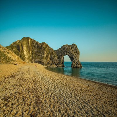 Durdle Door, an arch landscape formation, shows an arch in the rocks at the end of a small coastal sandy bay. The sun is low in the sky casting a soft light onto the rocks and the sea, which is very calm