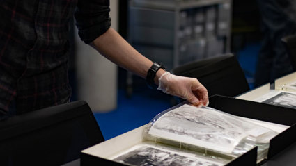 A person wearing archival gloves viewing historic black-and-white photographs stored in plastic sleeves and archival boxes.