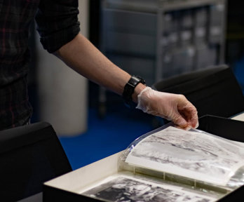 A person wearing archival gloves viewing historic black-and-white photographs stored in plastic sleeves and archival boxes.