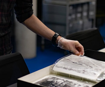 A person wearing archival gloves viewing historic black-and-white photographs stored in plastic sleeves and archival boxes.