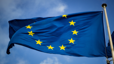 A blue with yellow stars configured in a circle, EU flag waves in the wind whist attached to a flag pole. The background is a partially cloudy blue sky.