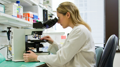 A person sits in a science labatory whilst looking through a microscope.