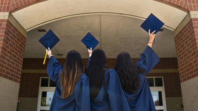 Three students stand before a university campus building in their graduation gowns. They are all holding their graduation caps in the air. 