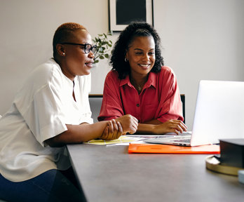 Two people sit at a large, modern table in a modern office setting, looking at a laptop screen.