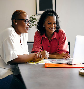 Two people sit at a large, modern table in a modern office setting, looking at a laptop screen.