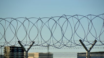 Close-up of a metal fence topped with spiraled barbed wire against a clear blue sky and blurred urban buildings in the background.