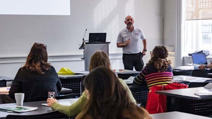 Group of people sitting at desks in a modern room listening to a presenter standing in the front.