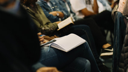 A row of people are seated, some with a notepad and pen on their laps. Others have their arms crossed or resting on their legs.