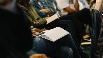 A row of people seated, some with a notepad and pen on their laps, others with their arms crossed or resting on their legs.