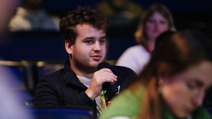 An audience member sitting in a dark lecture theatere at a conference, holding a microphone about to ask a question.