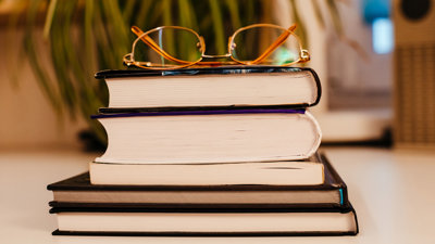 A pair of glasses rest on a stack of five books are stacked on top of each other.