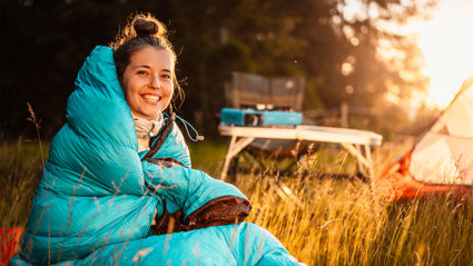 A person in a sleeping bag sitting in the outdoors next to a tent and foldable picknick table on a sunny day.