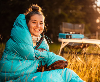 A person in a sleeping bag sitting in the outdoors next to a tent and foldable picknick table on a sunny day.