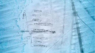Aerial view of a blue-tinged ice sheet with words in an Indigenous language etched in rows and faint tracks on the right.