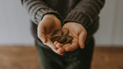 A person stand on a hard wood floor with there hands out and placed together, holding copper coins. 