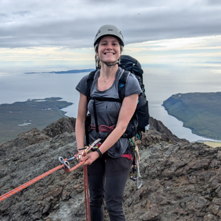 Kathryn Green posing for a photo. She is wearing climbing gear and is standing on the edge of a cliff overlooking a sea.