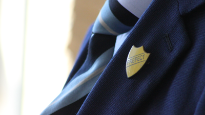 A secondary school student stands turned to the side, showing thier prefect badge.