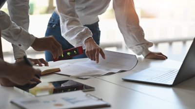 People reviewing architectural plans on a desk with a level, notebook, and laptop.