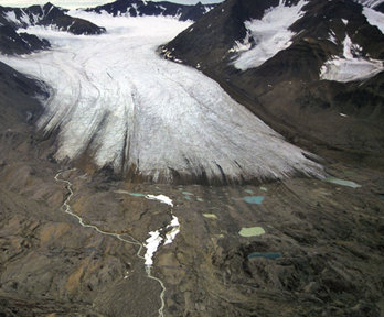 A glacier snout seen emerging from a valley. The glacier is white grey in colour and the valley sides are deep brown and scattered with snow fall