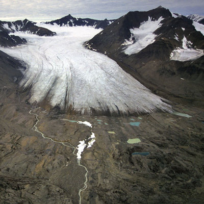 A glacier snout seen emerging from a valley. The glacier is white grey in colour and the valley sides are deep brown and scattered with snow fall