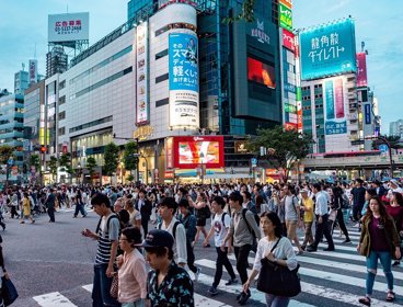A very busy road crossing in Tokyo, with many people crossing the road at the same time. Zebra crossings can be seen, as well as large buildings with neon lighting in the background