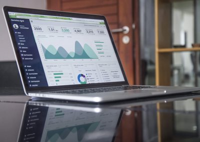 A laptop sits on a glass table with an analytics website presention ont he screen. graphs. charts and various numbers are shown. labels sre not legible.