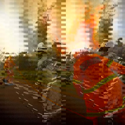Two firemen in orange uniforms fight a fire by the side of the road. The flames are reaching high into the air and there is lots of smoke