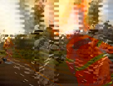 Two firemen in orange uniforms fight a fire by the side of the road. The flames are reaching high into the air and there is lots of smoke