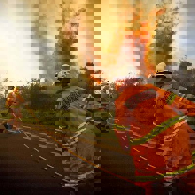 Two firemen in orange uniforms fight a fire by the side of the road. The flames are reaching high into the air and there is lots of smoke