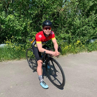 Nick Hart on a racing bicycle on an asphalt road and in front of a bush.