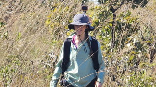Researcher walks through tall pale brown grass and small trees wearing a rucksack with a LiDAR scanner attached.