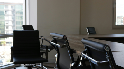 Black conference chairs sit around a square wooden table. One of the chairs sits away from the table unlike the rest