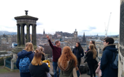 A group of people listening to a speaker on Calton Hill. The background shows the Dugald Stewart Monument and the Edinburgh skyline.