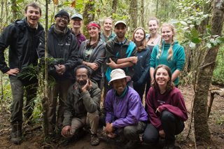 A group of nine people standing and three people crouching in a forested area.