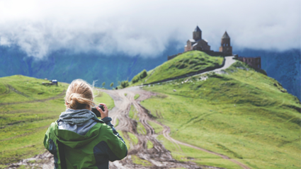 A person stand on a path leading up to a castle on a grassy hill whilst looking into their camera and taking a picture