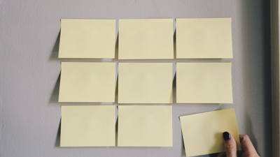 Seven plain yellow post it notes are configured in to a square with the right corner incomplete. A hand with black nail polish is in the midst of placing the eighth plain yellow post it note to fill the gap in the square