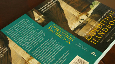 A book titled 'the royal geographical society expedition handbook' lays open but face down on a wooden surface.