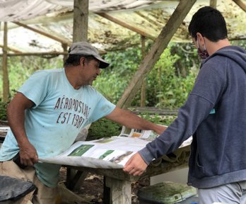 Two people under a planting shelter holding out a large piece of paper on a wooden table. The two people are looking and pointing at the paper. 