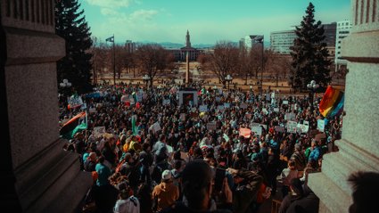 Large crowd gathered in a city park for a peaceful protest on a sunny day with banners and flags visible.