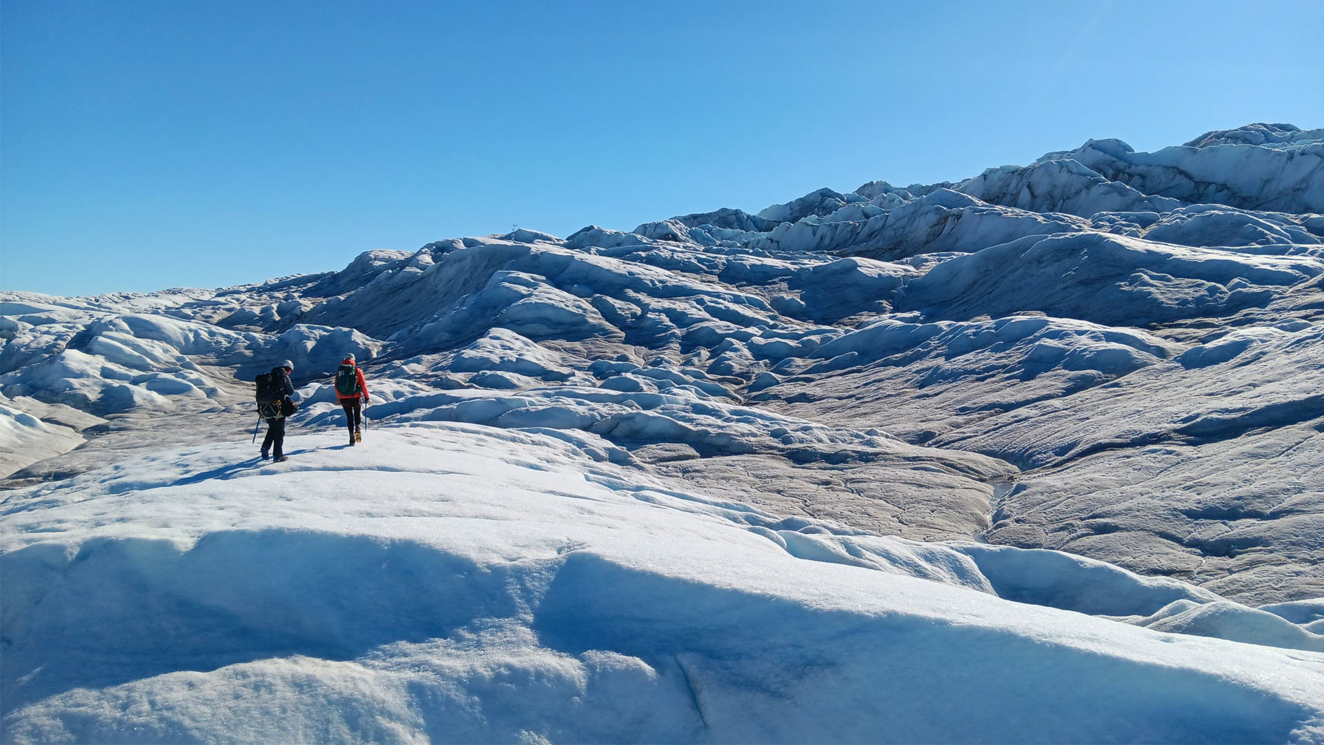 Two trekkers on a glacier crossing a rugged ice sheet on a bright, sunny day.