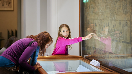 A person hunched over a display cabinet and a young child pointing at an old world map.