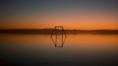 A children's swing in the middle of a lake, an orange sky reflecting on the water's surface.