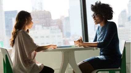 Two people sit across from each other at a small white table. They appear to be mid conversation with their hands resting on the table. 