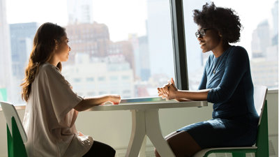 Two people sit across from each other at a small white table. They appear to be mid conversation with their hands resting on the table. 