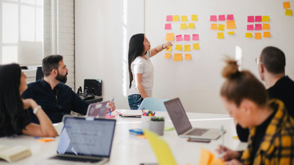 People sat around a table typing on laptops. One person is stood at the front pointing at square sticky notes stuck on a whiteboard.